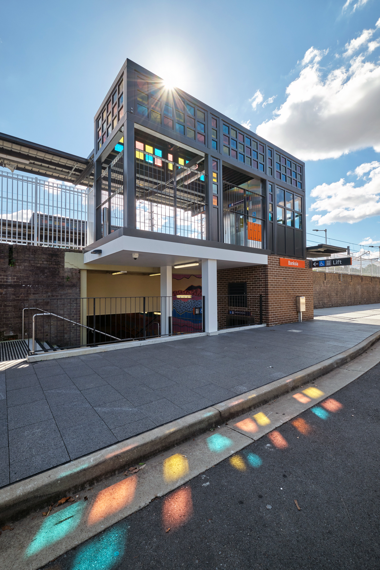 The roof providing weather protection outside the platform level of the lift features squares of coloured glass. The sun is shining through the glass creating coloured patterns on the road below