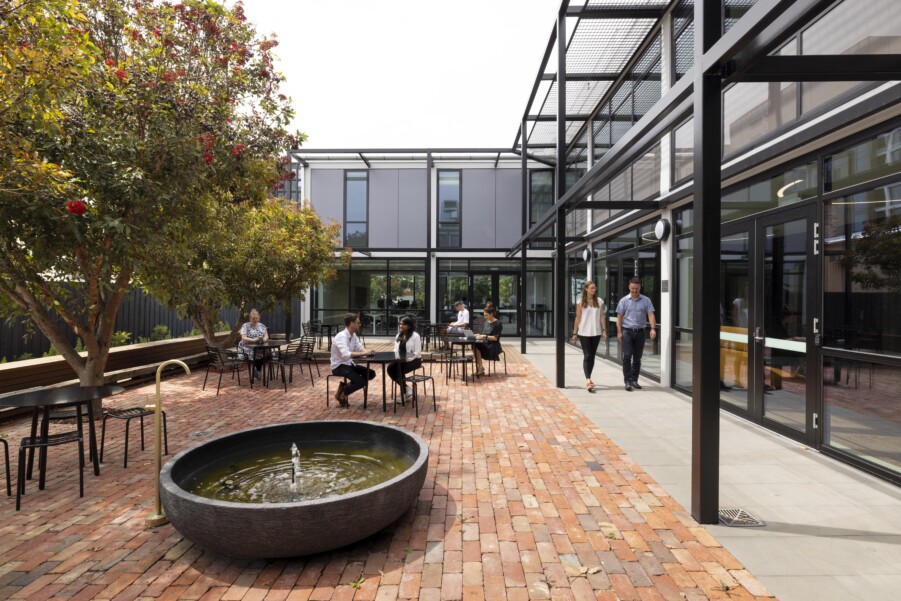 An outdoor area with trees, brick pavement and a small fountain, few people are chatting and working in the seating area.