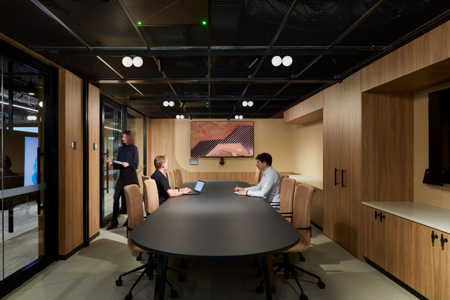 The formal boardroom style meeting space has a large matt black table at the centre. Employees sit at high-backed natural leather chairs working together on a laptop and a large display screen showing solar panels. Another staff member has come into the room and is closing a sliding glass door to the space. The outer office is visible through the full height windows and doors separating the space. White globes of light 'float' below the steel framing of the ceiling.