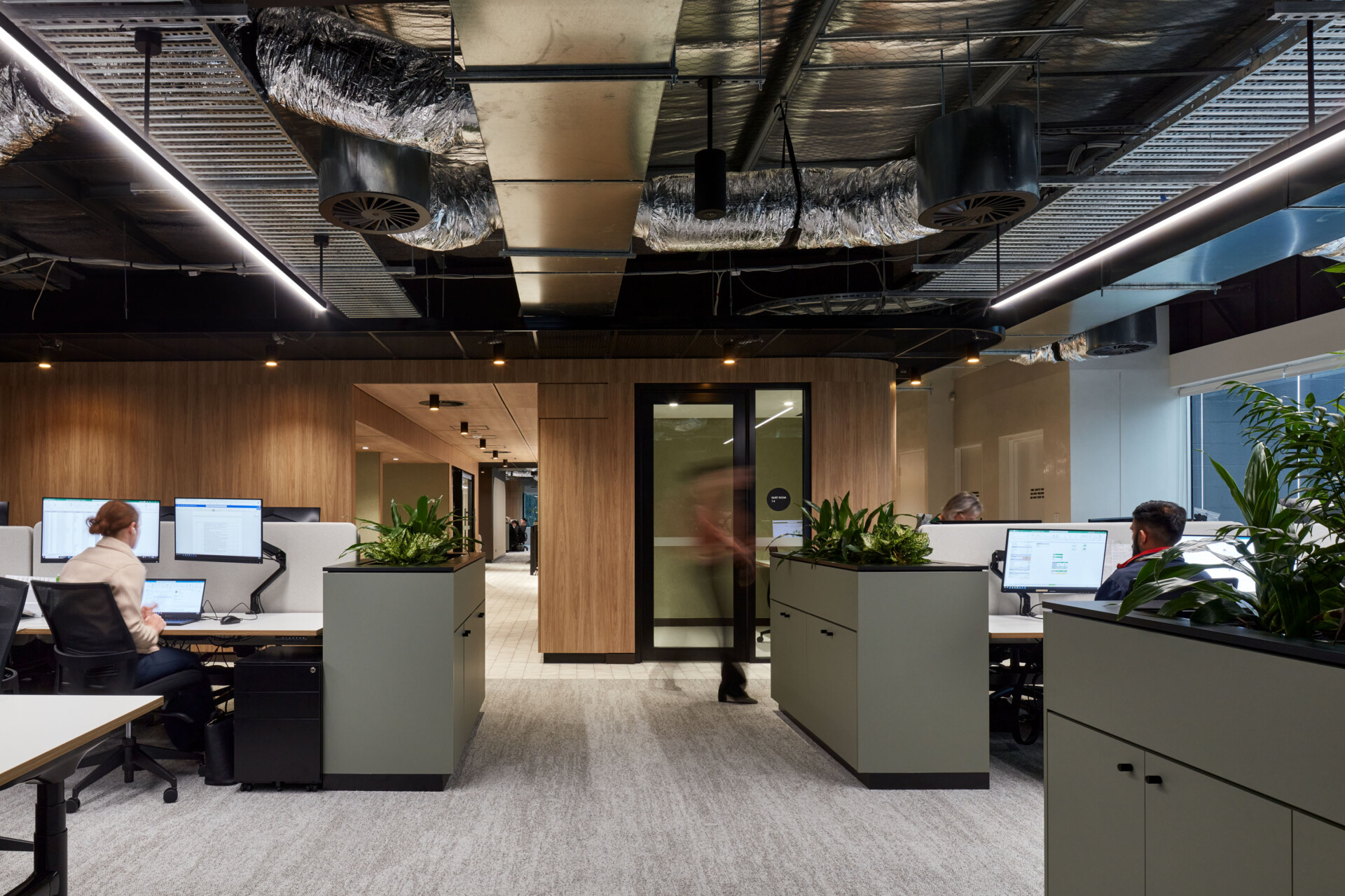 One of the open plan workspaces. Staff sit at standing and sitting desks working in front of screen. You can peek into a quiet room adjoining the workspace through its glazed door and sidelight. A corridor leads to the other end of the floor with a peak into work booths along its length. Planter boxes with oxygen purifying plants top each work bank.