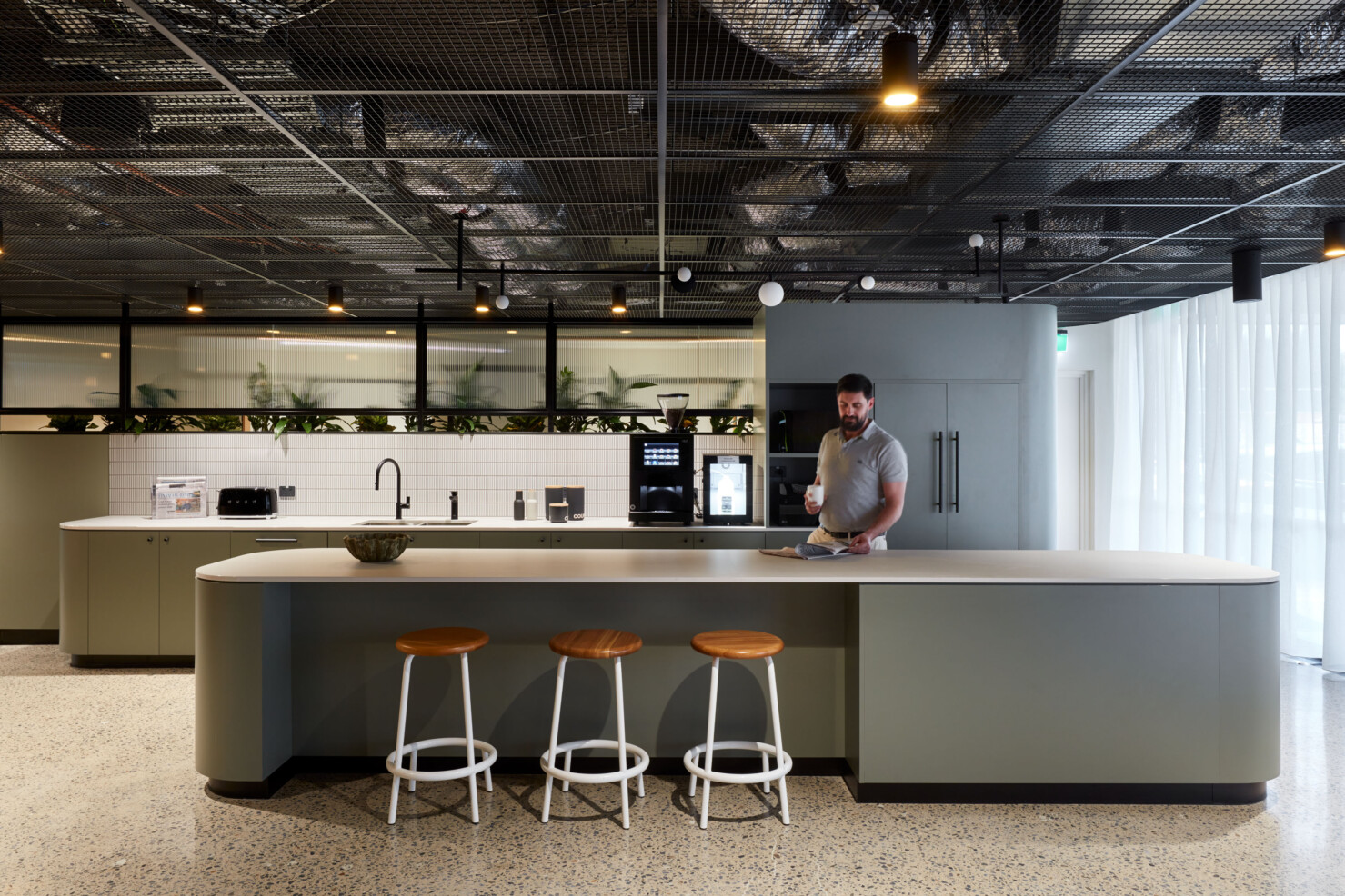 A man reads the paper while drinking a cup of coffee in the kitchen. White bar stools with warm timber tops sit under an island bench in pale green with a white top. White tile splashback and matt black tapwear feature. Appliances are hidden within the pale green joinery that separates the kitchen from the lockers and amenities. Natural light filters through privacy curtains and the full height windows that lead to the staff carpark.