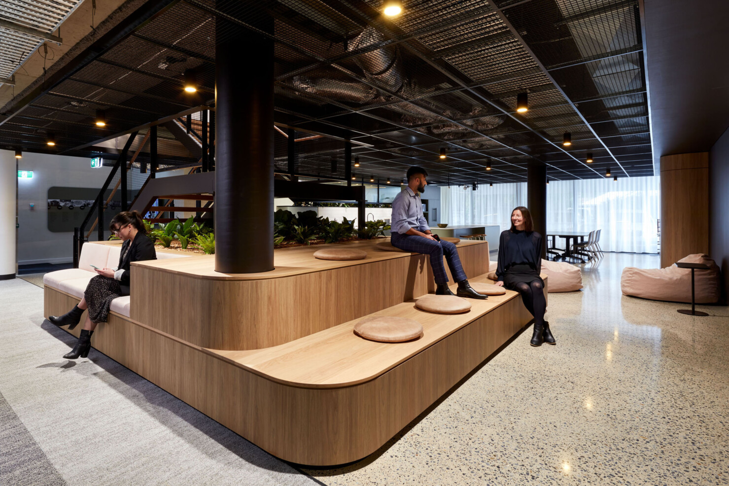 A central feature stairway becomes the gathering point for staff to move about the office and interact with each other. Polished floors, black steel and exposed services are softened by plants, custom joinery in warm wood and soft furnishings in pale pink. A floating platform provides a tiered seating space for staff to sit or visitors to wait near the entry of the office. A large kitchen and dining area is in the background with sheer curtains letting in light from the full height windows to the rear of the space.
