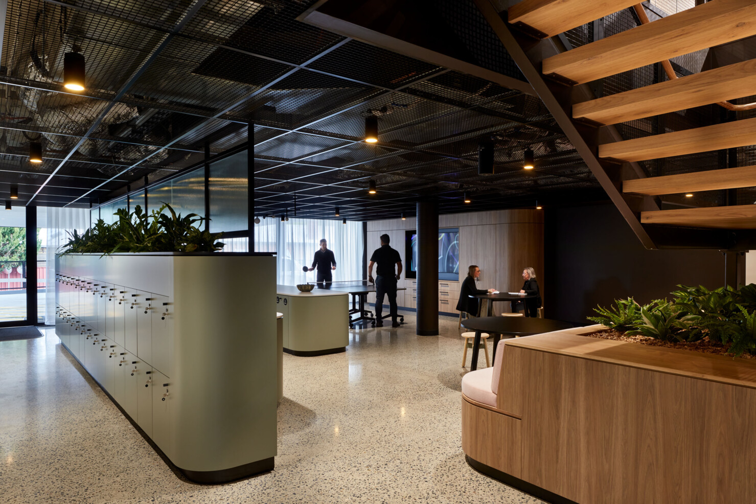 A central feature stairway becomes the gathering point for recreation and for staff to move about the office and interact with each other. Polished floors, black steel and exposed services are softened by plants, custom joinery in warm wood and soft furnishings in pale pink. A floating platform provides seating space together with cafe-style furniture and beanbags. A large kitchen and dining area is in the background with sheer curtains letting in light from the full height windows to the rear of the space. A large kitchen and breakout space on the lower level provides a multi-functional space for staff to interact and industry partners to gather together. Two staff play table tennis on the table in the breakout space. Another couple of staff hold an informal 'cafe style' meeting on a round black table in the space. A bank of lockers create the backbone of the kitchen and separate the space from amenities and are located near the car park for ease of access.