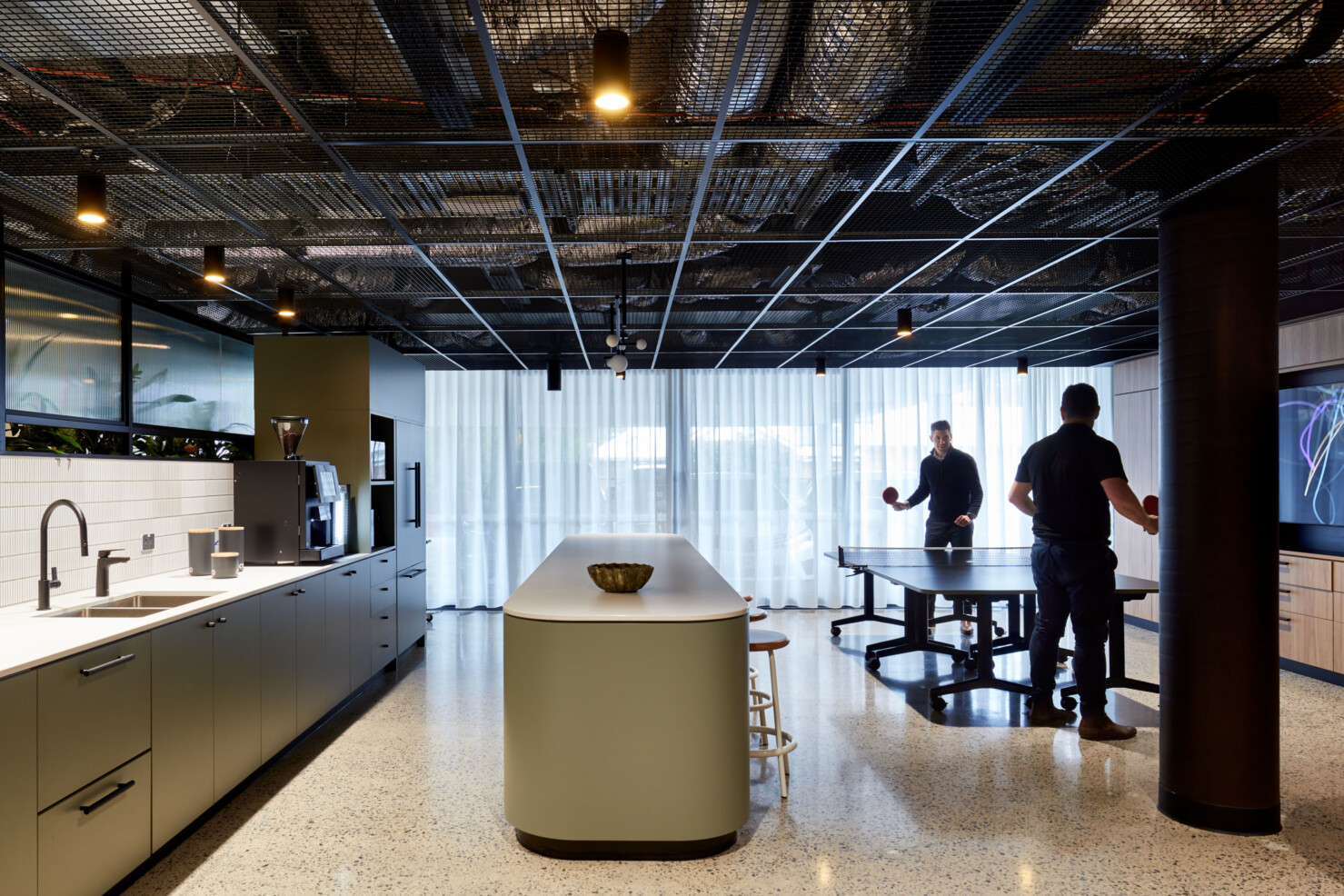 The lower level kitchen and staff breakout area has polished concrete floors, built in joinery housing fridges and other appliances. Modern fittings and fixtures in matt black suit the industrial aesthetic of the building interior and exposed steel structures. An island bar in rounded pale green divides the space. Two staff play table tennis on the large dining table.