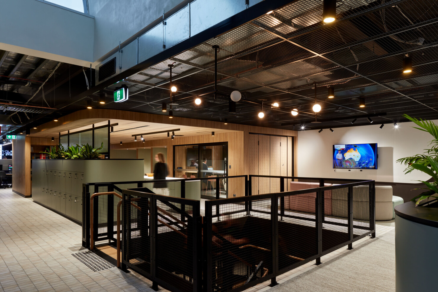 A central feature stairway connects the lower and upper floors and provides a focal point of the communal zones. Level one of LMS Energy Headquarters arrives at a communal space with modular lounges in sand and pale pink. Sandy coloured tiles lead through a breakout kitchen in pale green with matt black fittings and fixtures, and white benches. A staff member walks past the stairway and two staff meet in a meeting room in the background. A bank of lockers run the length of back of joinery that holds a tea point and appliances. The warm timber joinery softens the black steel framing that separates the exposed services in the ceiling space.