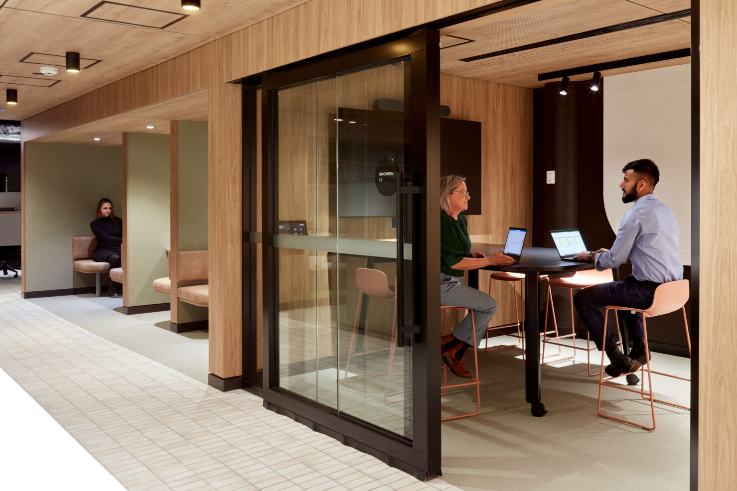 Two staff with laptops sit on light pink chairs at a matt black worktop in a small meeting room. A black screen is on the wall at the end of the bench. A large custom-designed whiteboard is visible on the back wall. The warm timber joinery creates individual rooms and booths. Another staff member sits working in a quiet booth in the background.