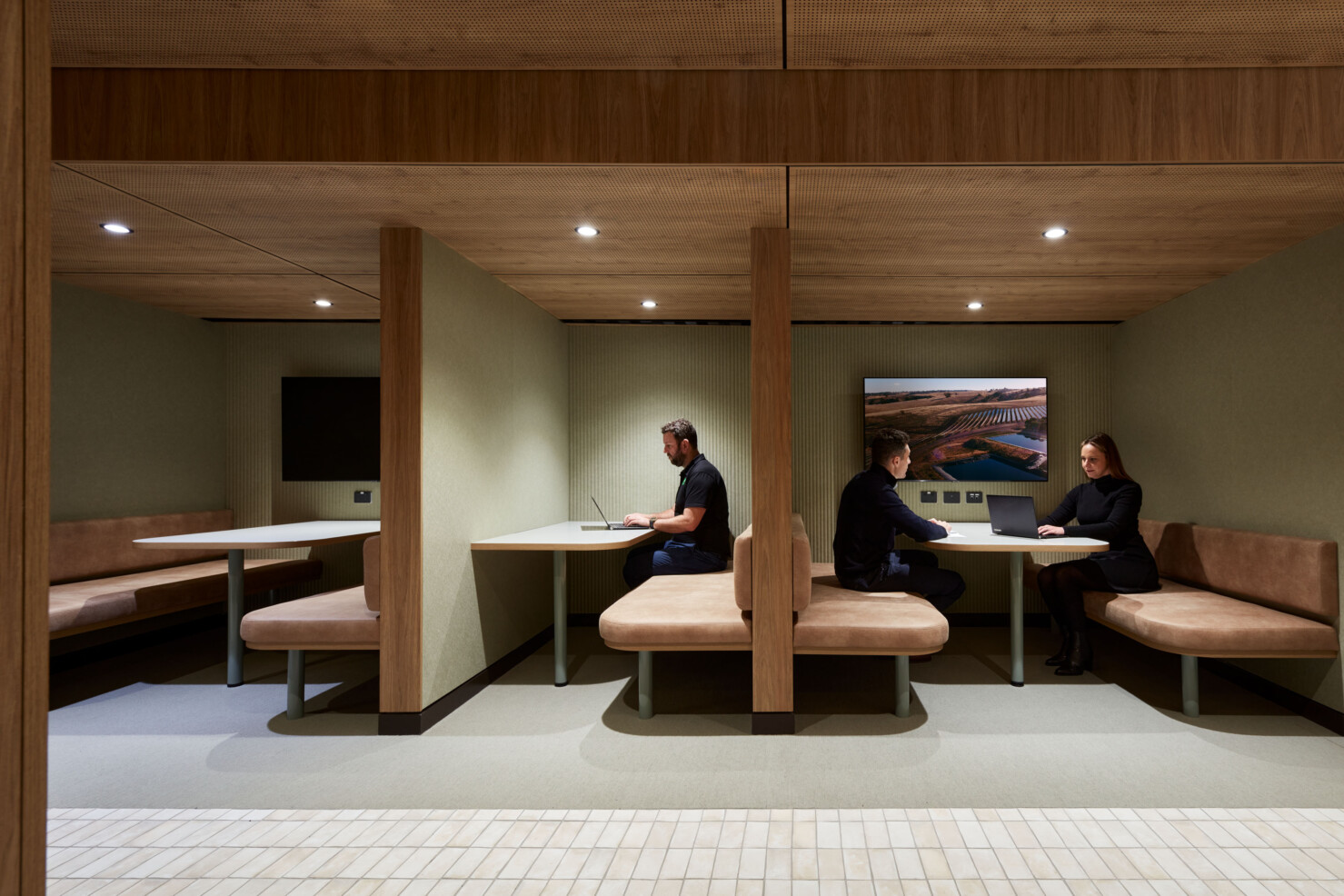 Two staff sit in a quiet booth working on a laptop connected to a screen on the wall. A picture of solar panels in a landscape is on the screen. Another staff member sits solo in an adjoining booth working on a laptop. The booths feature warm timber joinery, sage green sound-absorbing wall lining and natural-coloured leather seat pads.