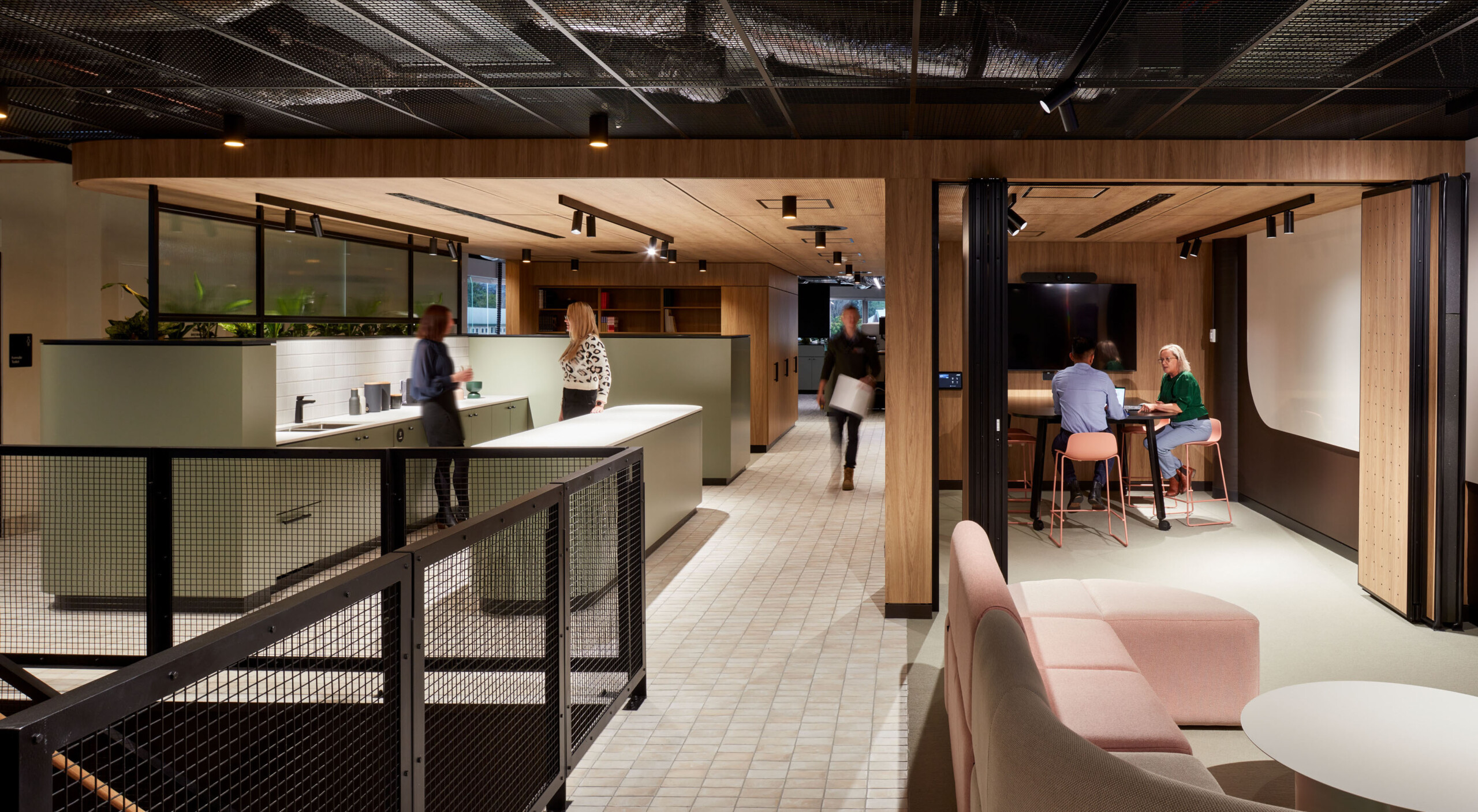 Level one of LMS Energy Headquarters arrives at a communal space with modular lounges in sand and pale pink. Sandy coloured tiles lead through a breakout kitchen in pale green, matt black fittings and fixtures, and white benches. A couple of staff gather in the kitchen chatting. Another staff member walks through from the offices at the rear with a box. Two staff meet in a meeting room off to the side that has concertina doors that open to the lounge for larger groups to gather. The warm timber joinery softens the black steel framing that separates the exposed services in the ceiling space.