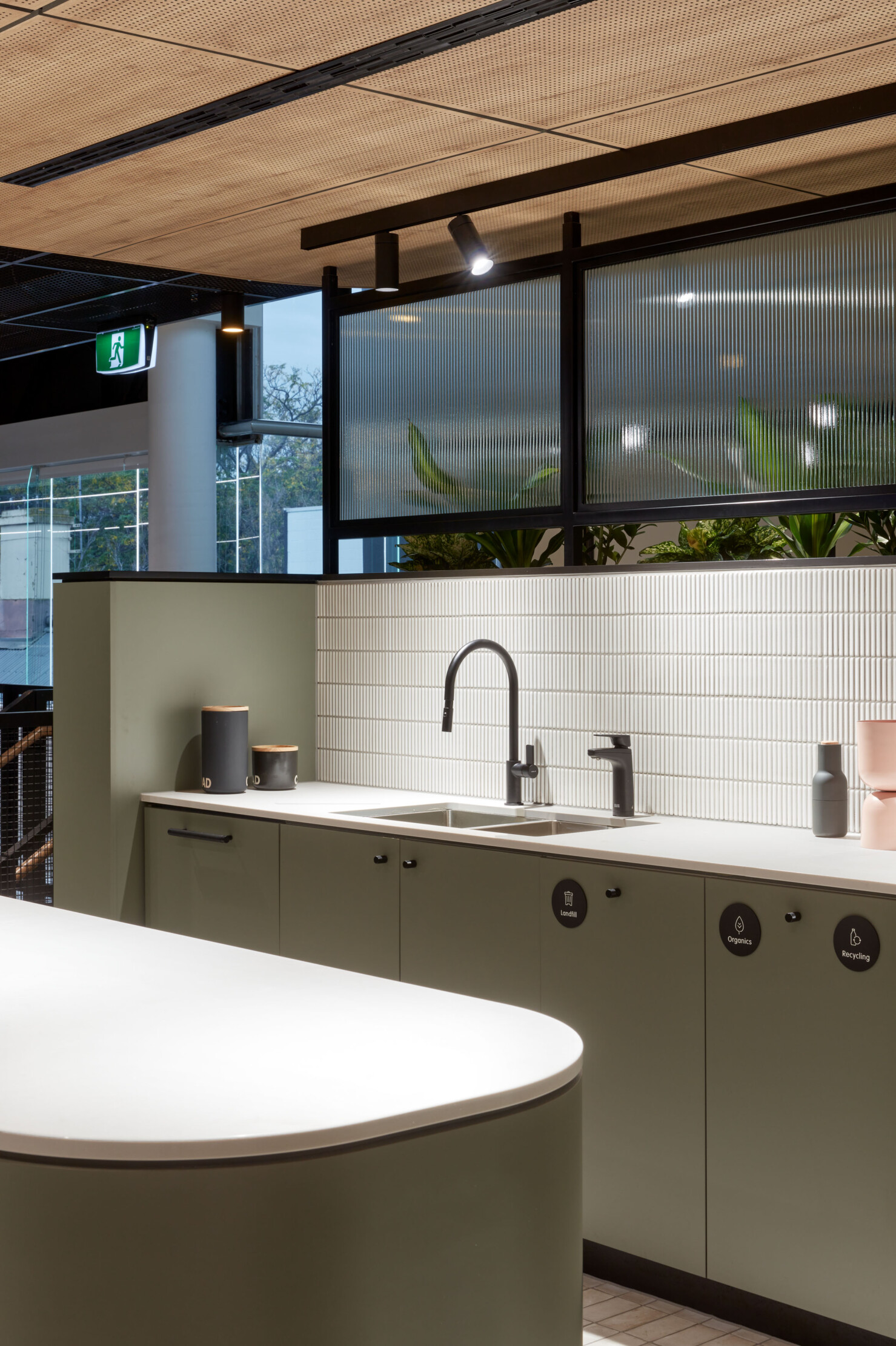 A tea point on the upper level provides a central kitchen for food preparation featuring matt black tapware and white tile splashback. The appliances and waste disposal systems are all hidden away behind pale green joinery. A planter box and glazed screen separate the space from the amenities.