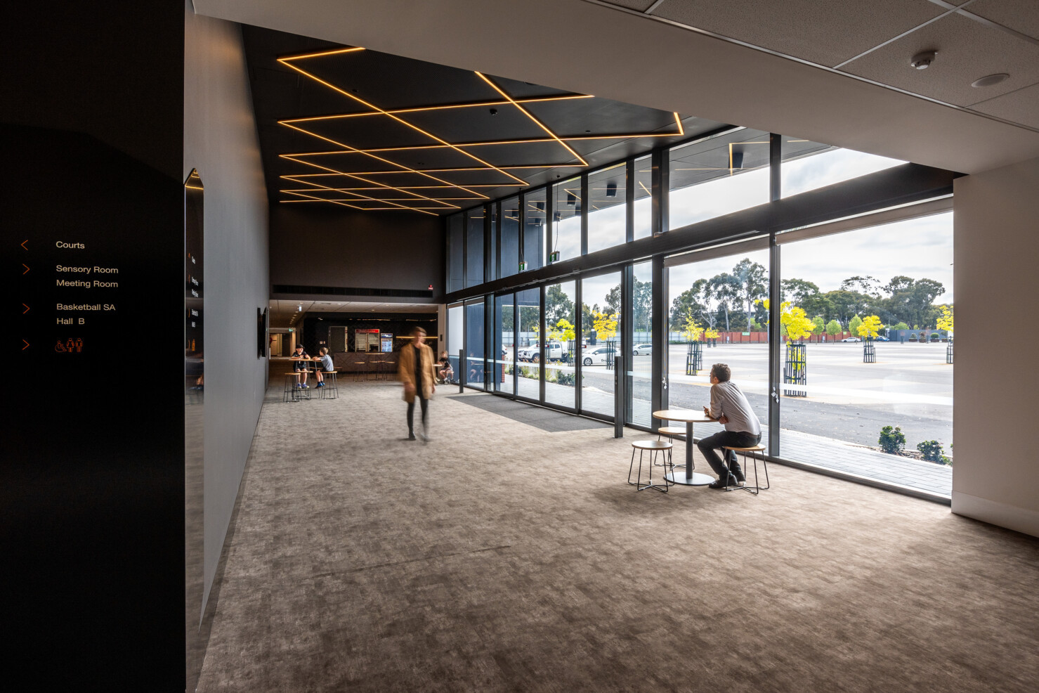 The entry and spectator foyer features a criss cross of LED strip lighting that replicates a basketball net. People wait in the foyer, walking the space and sitting on cafe tables.