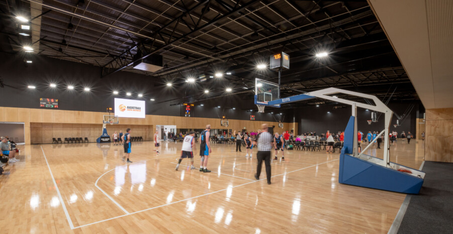 The stadium has a black canopy with light ply cladding on the walls at the court level. Basketball teams play a practice match on the high gloss maple court with an umpire in the foreground watching the game.