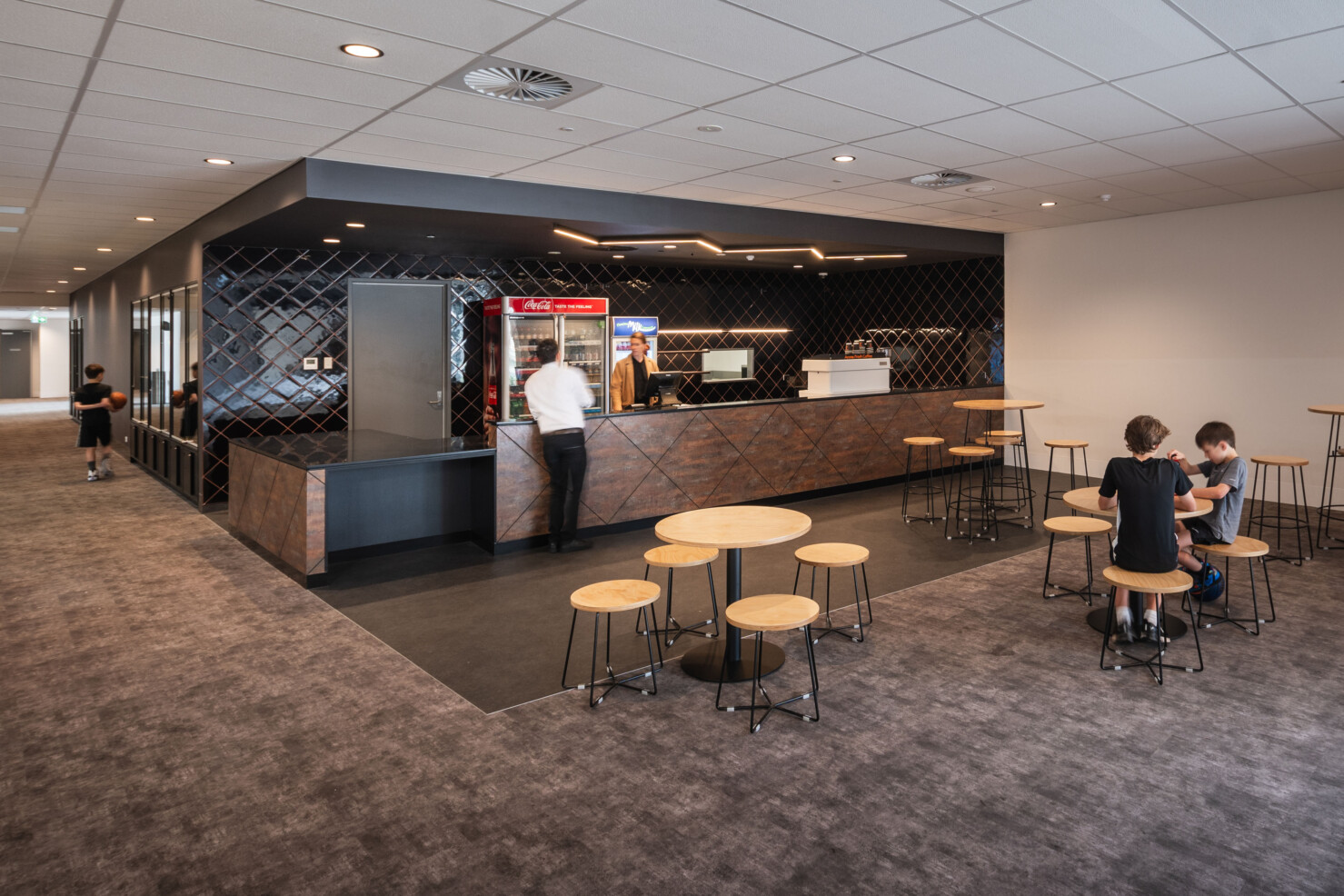 The cafe and bar in the spectator foyer provides seating and refreshments for the public. A person serves a customer from the counter. Two young boys sit at the cafe tables enjoying a soft drink. Another young person walks up the corridor holding a basketball. The bar features black gloss tiling.