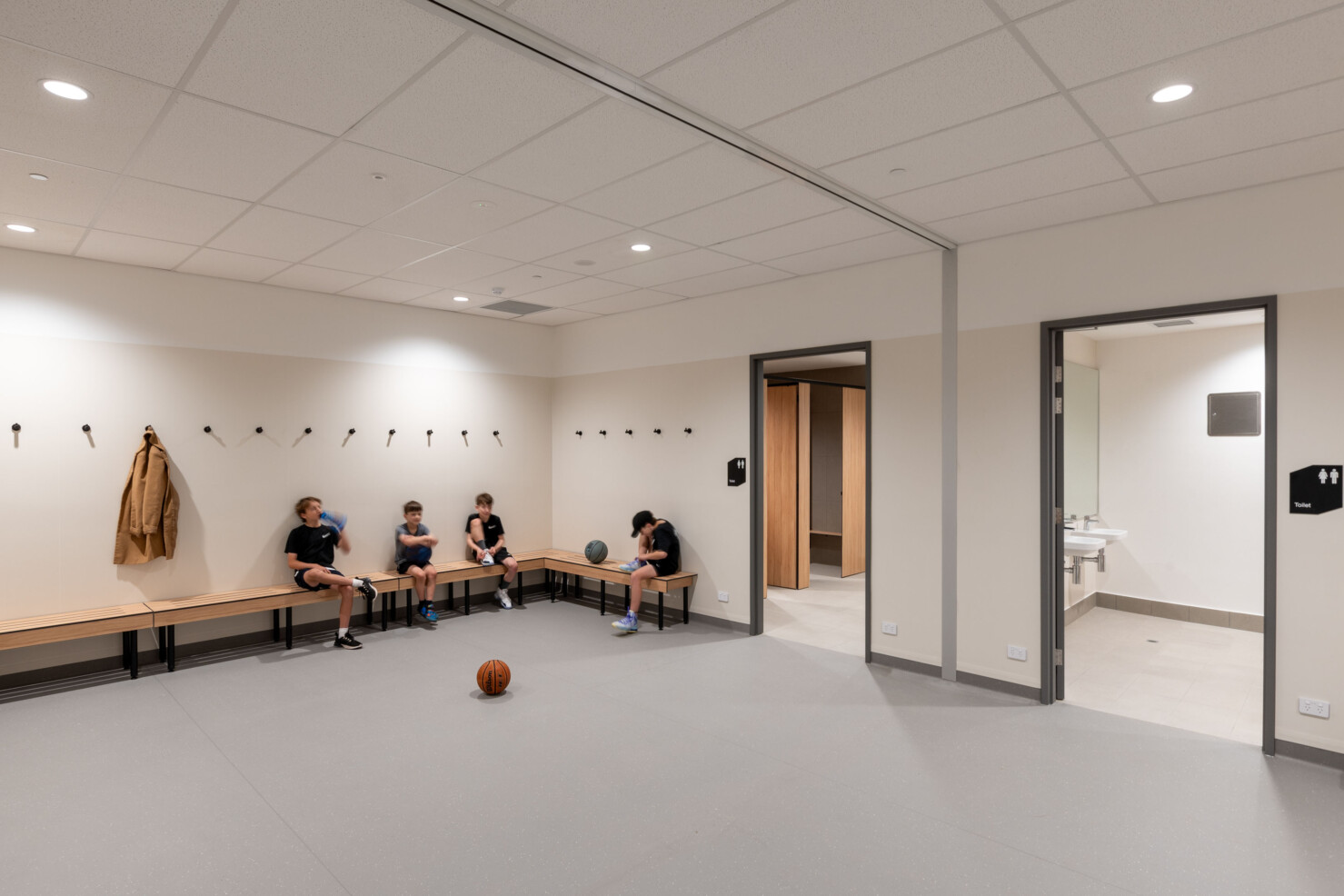 Clean and minimal change facilities provide ample space for accessibility. Kids sit with their basketballs, talking and drinking from water bottles on the light coloured timber benches.
