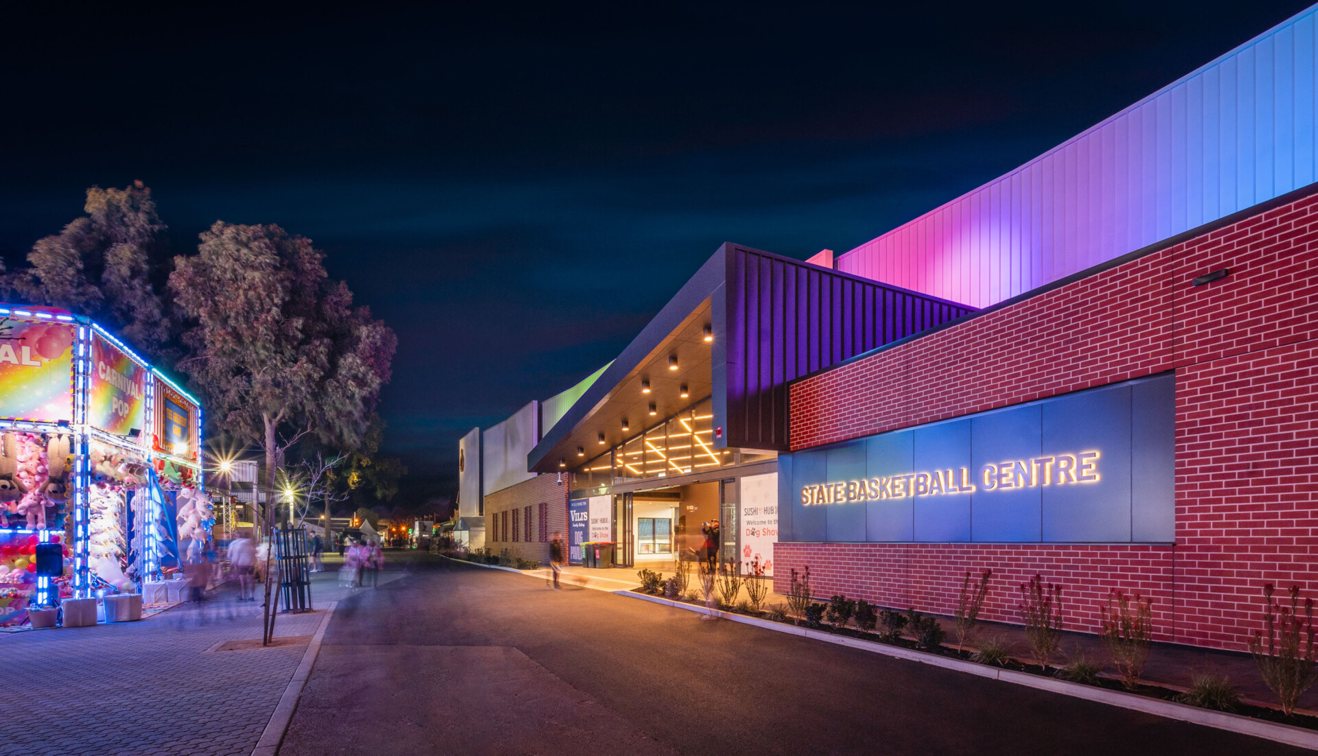 A carnival spirit is in this shot at night. Showground sideshows are installed adjacent to the building which is lit up in pinks, purples, blues and green light projected on the canopy. The building name is LED backlit and glows warm white together with the cross hatched lighting of the entry foyer.