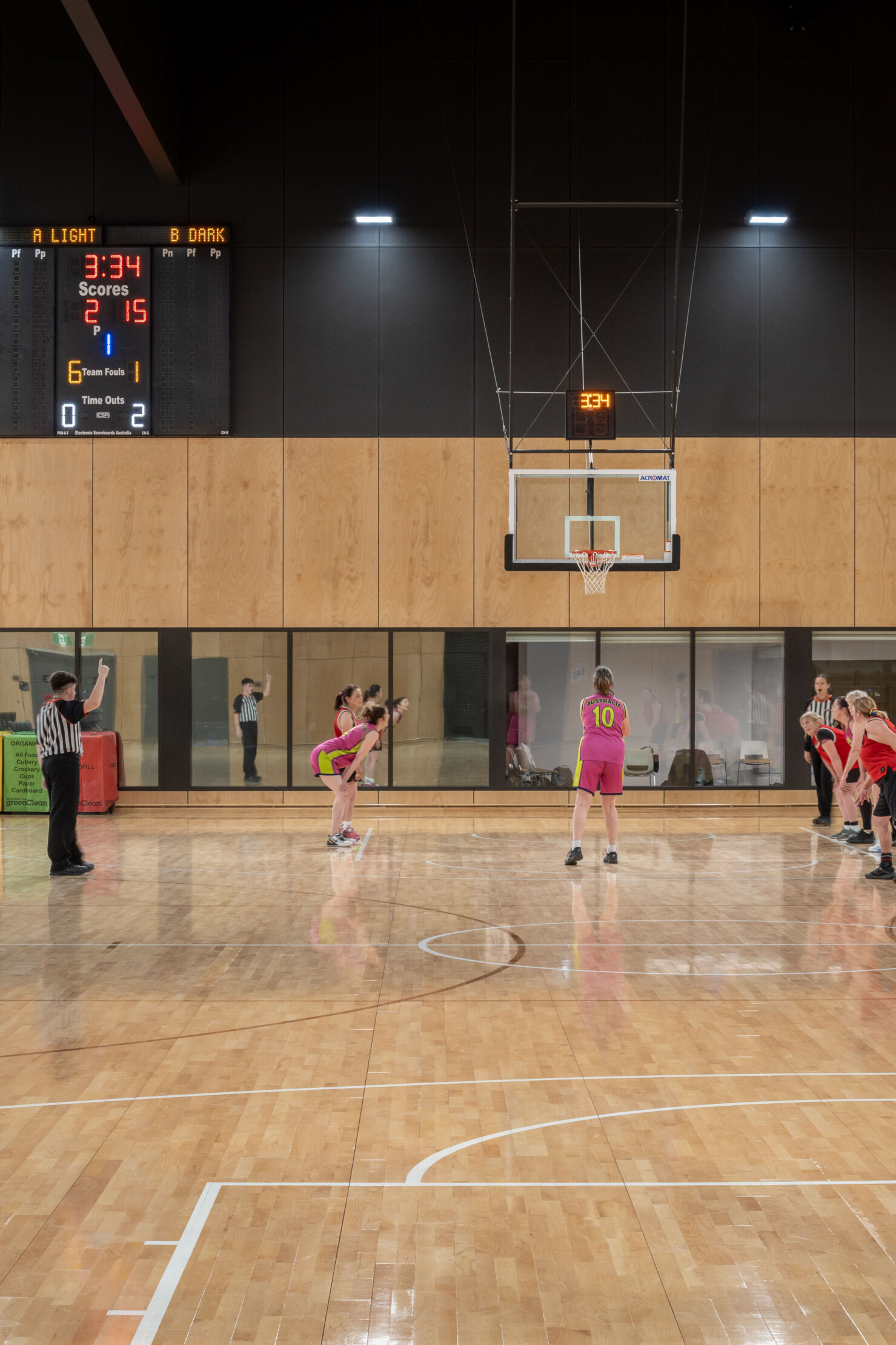 Female basketballers in pink uniforms crouch at the goals as a penalty shot is thrown by a player. An umpire stands by refereeing the match on the maple lined courts.