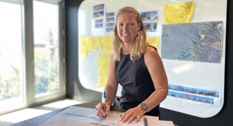 Design Manager, Kat Povey, is seated at a work bench with schedules, a notebook. In the background are site plans, architectural drawings on a big whiteboard on the wall. There is natural light filtering through the full height window on the left.