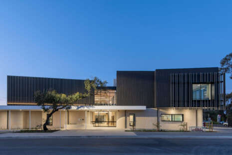 Tiwu Kumangka at dusk. The warm sandstone blockwork and dark timber-look upper level glows with architectural lighting. The building name is black above the entry canopy. A gum tree reaches accross the front entryway. Strong vertical fins wrap the upper level.